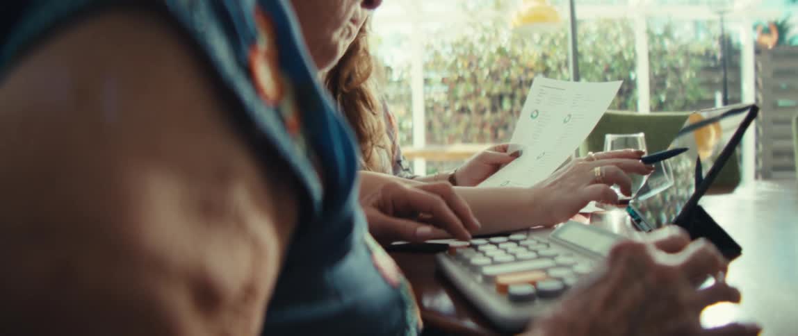 A view from the side shows an older woman and a younger woman sitting at a table together while they review financial documents and use a calculator and a tablet