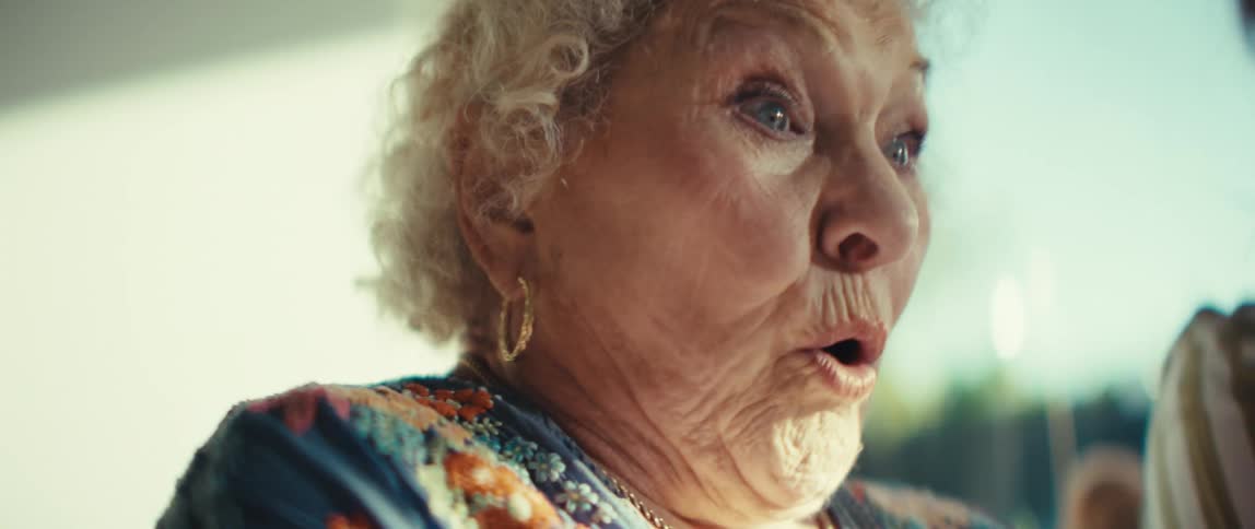 A close view of an elderly woman with white curly hair as she looks at a tablet screen and reacts with a surprised facial expression while sitting next to a younger woman in a bright room