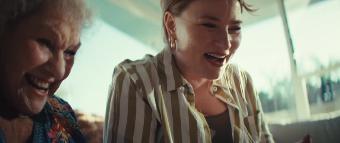 An older woman and a younger woman sit closely together laughing and smiling while holding video game controllersthe scene captures a joyful multigenerational bonding moment as they play a game indoors