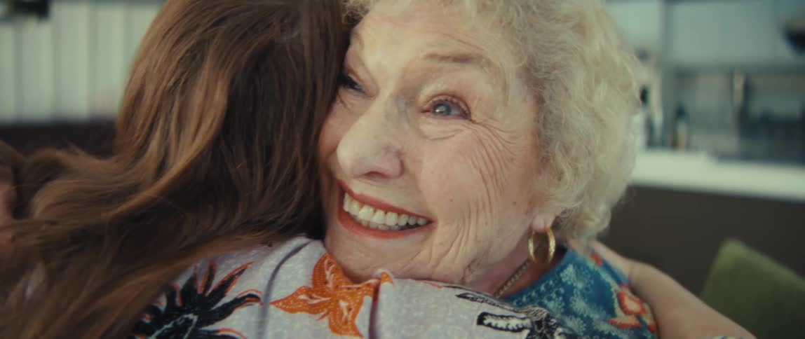 An elderly woman smiles as she warmly hugs a younger woman in a bright cozy kitchen setting