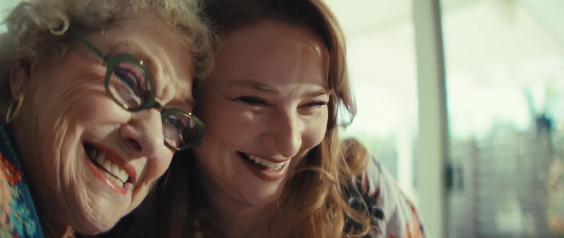 A younger woman with long red hair and an older woman with glasses smile and laugh together while looking at a tablet on a table inside a bright home