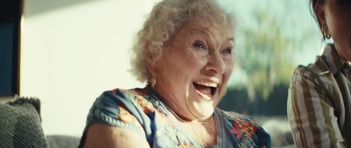 An elderly woman with curly white hair laughs joyfully while playing a video game she holds a black game controller in her hands her face lit up with excitement and surprise