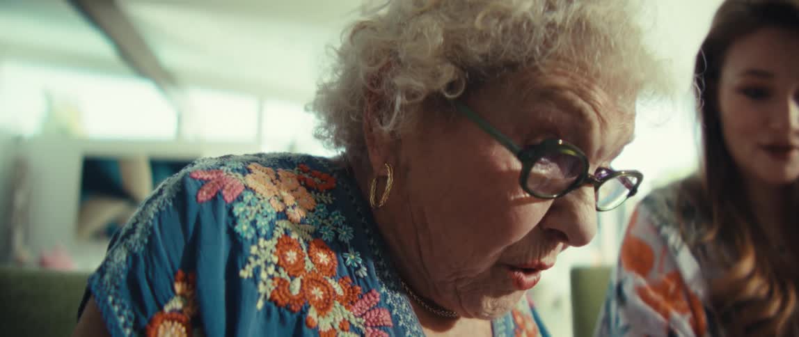 An elderly woman wearing glasses punching numbers on a calculator in excitement while a young woman beside her goes through a document both become excited in the living room at midday