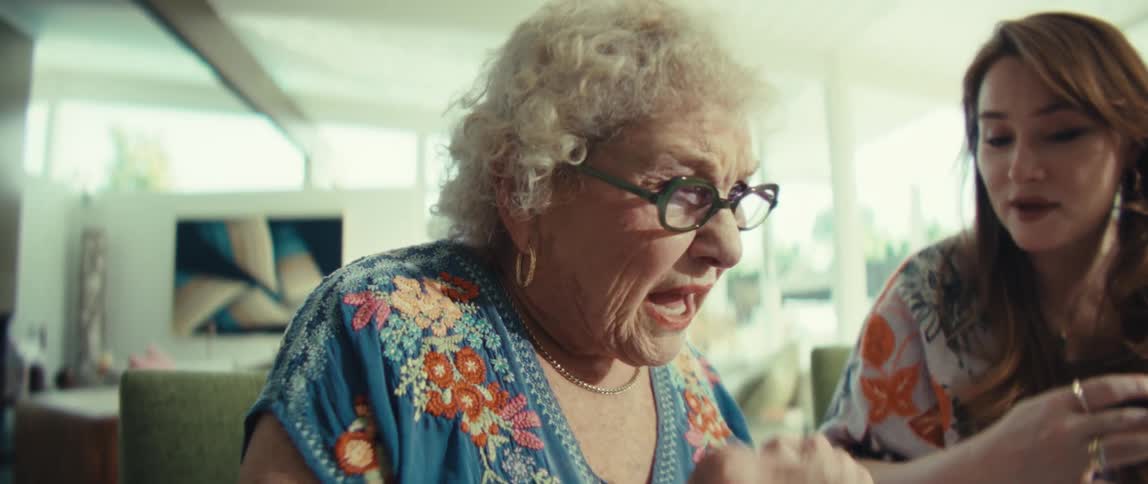 A younger woman assists an elderly woman who is using a calculator while they sit together at a table the scene captures a focused multigenerational interaction as the younger woman looks on and provides guidance with a tablet nearby