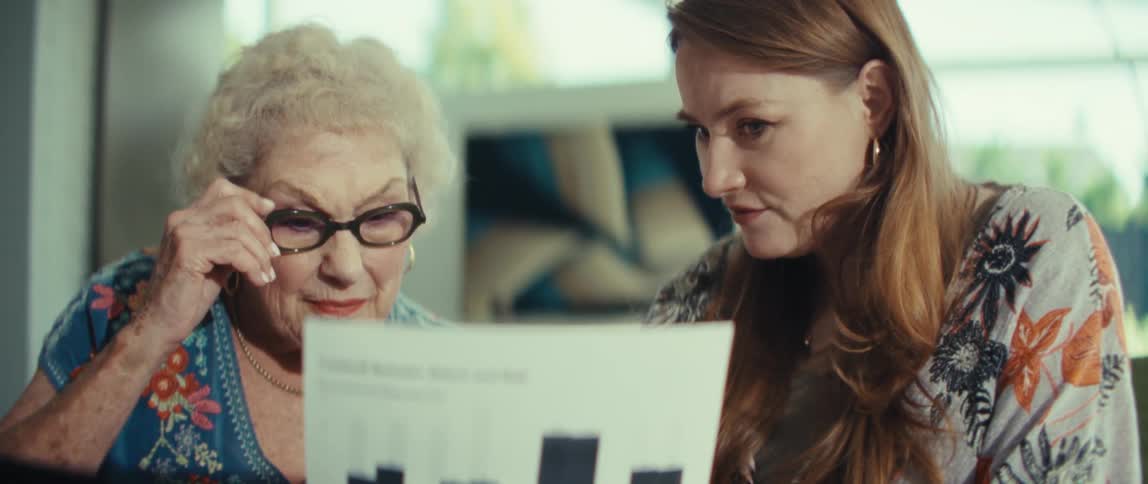 An elderly woman and a young woman sitting in front of a computer with the young woman holding a document and discussing it with the elderly woman in the living room at midday
