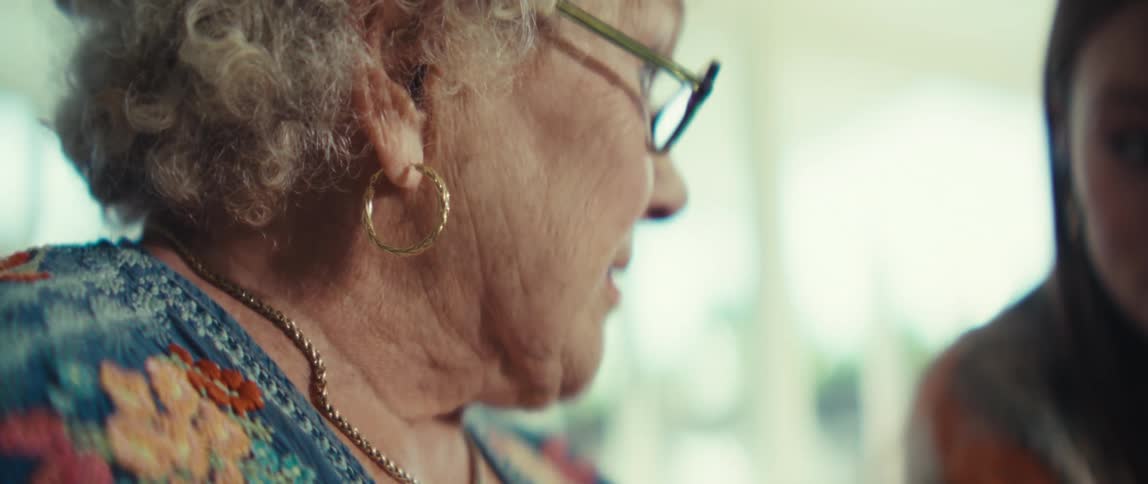A close view shows an elderly woman with white curly hair and glasses talking and smiling as she sits next to a younger woman who is also laughing in a bright room