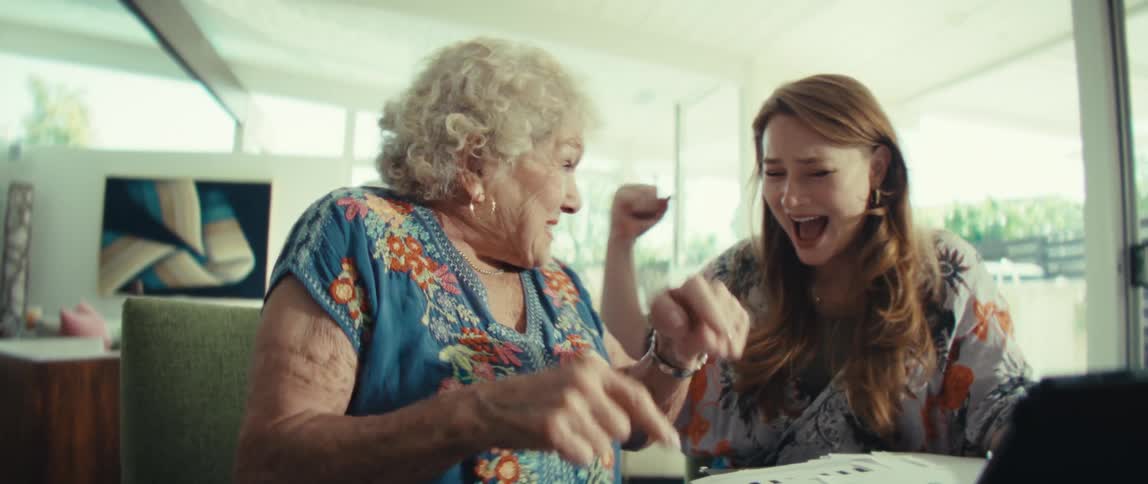 An elderly woman gasps in delighted surprise as she presses buttons on a calculator while a younger woman beside her smiles and points at a tablet sharing an animated joyful moment at the table