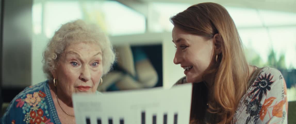 An elderly woman and a younger woman sit together looking at a paper with financial charts and they talk about the data before smiling and leaning toward each other in a bright indoor room with large windows behind them