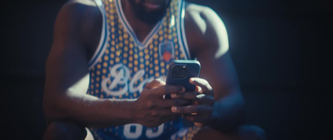 A young man sitting outdoors under the sun scrolling on his phone while wearing a sports jersey