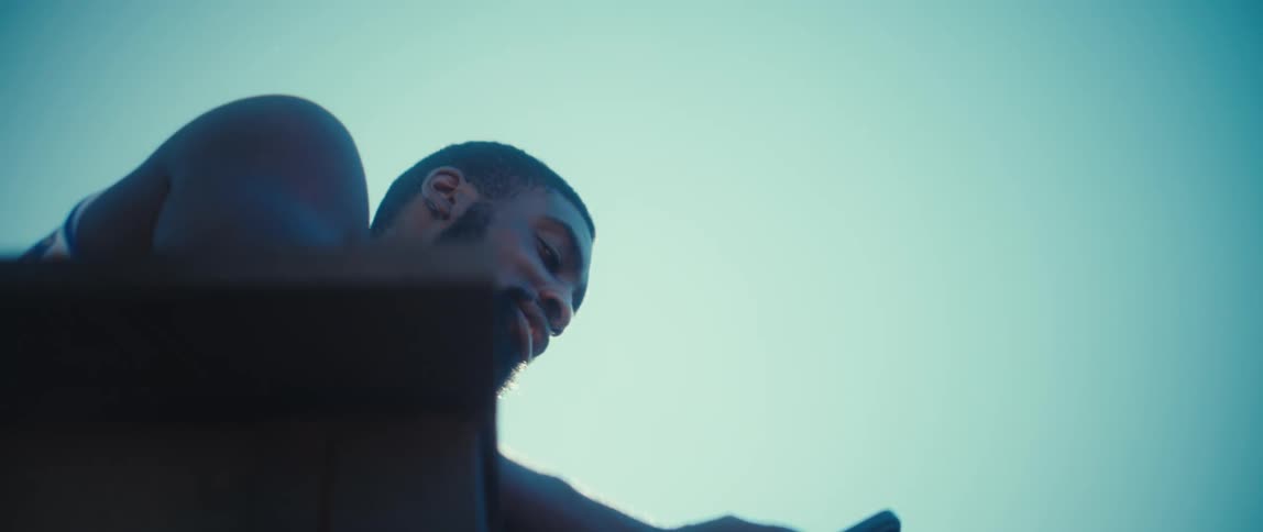 A black man stands on a balcony against a bright blue sky while looking down at a smartphone in his hands