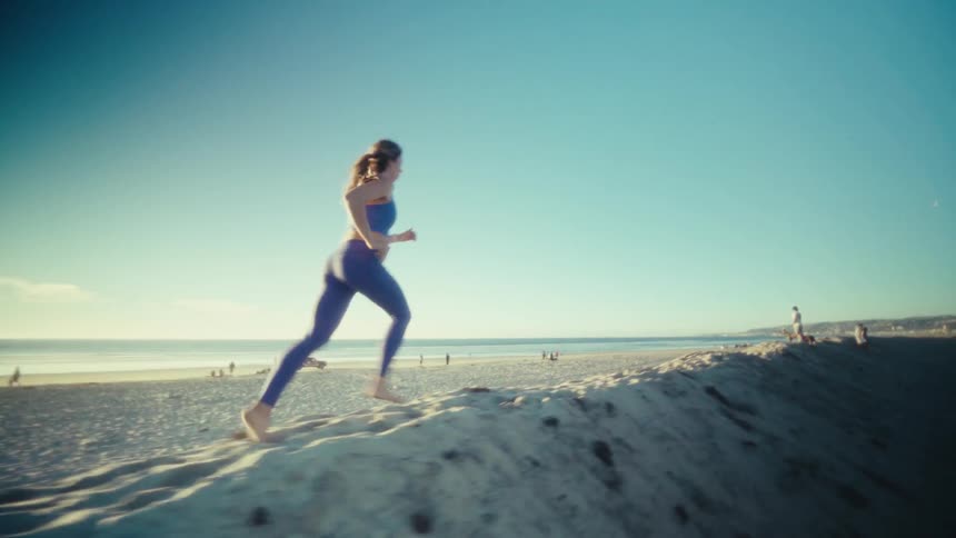 A view from a distance shows a pregnant woman wearing a blue sports bra and blue leggings as she runs across a sandy beach with the ocean and a pier in the background during sunset