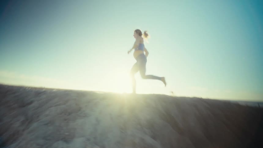 A pregnant woman in athletic wear runs along a sandy ridge during a bright sunny day the camera follows her movement at a low angle capturing the lens flare from the sun and the beachgoers in the background