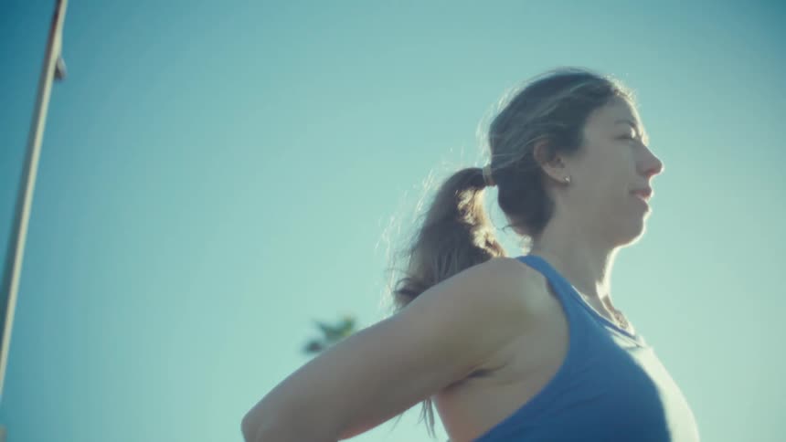 A pregnant woman wearing a blue sports top and leggings stands outdoors and stretches her leg while looking toward the horizon on a clear sunny day with palm trees in the background