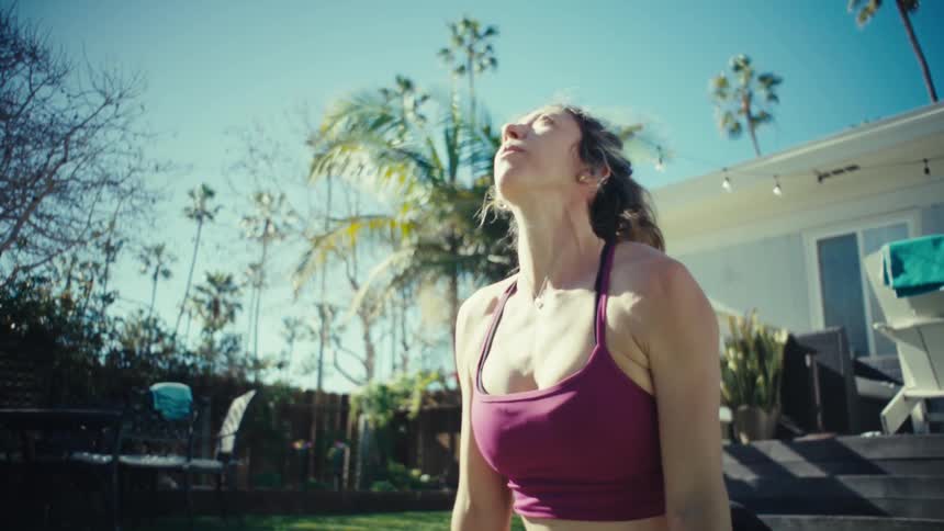 A woman wearing a maroon sports bra and black leggings performs yoga on a mat in a sunny backyard by transitioning from a plank pose into a downward dog pose while surrounded by green grass and palm trees