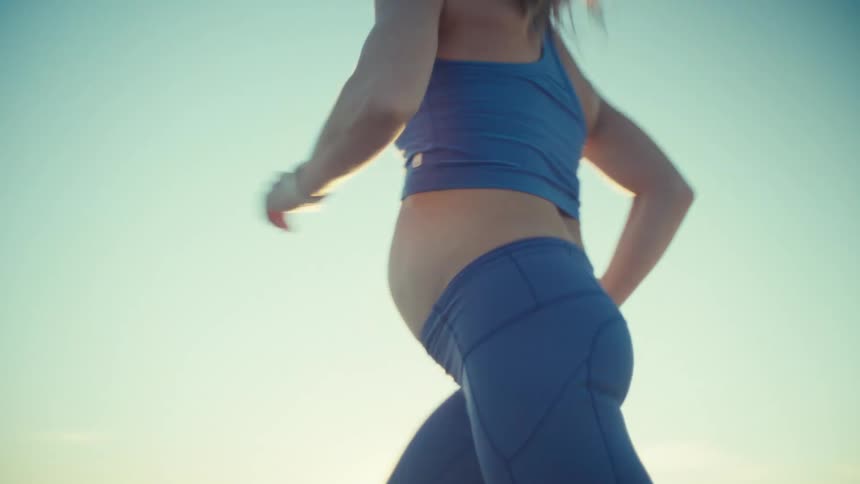 A pregnant woman in blue athletic wear runs along a sandy ridge at the beach during sunsetthe camera follows her movement as she jogs past capturing the warm light and the ocean in the background
