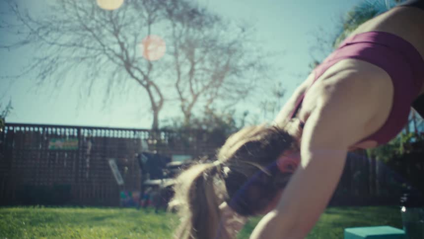 A pregnant woman wearing a maroon sports bra and black leggings performs yoga poses on a mat in a sunny backyard with green grass and trees