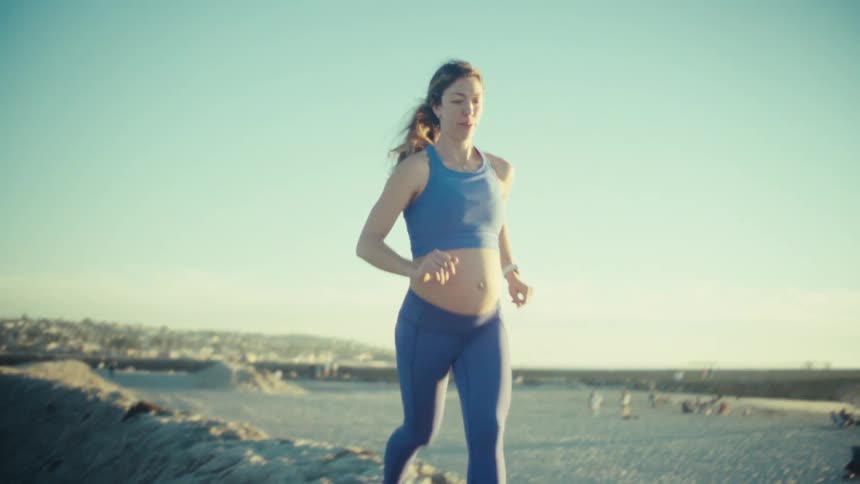 A view from the front shows a pregnant woman wearing a blue sports bra and blue leggings as she runs along a sandy ridge at a beach during sunset