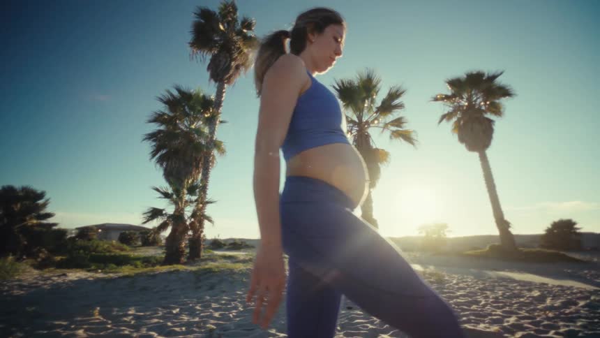 A pregnant woman in a blue athletic top and leggings walks across a sandy beach during a bright sunny day she moves past tall palm trees as the sun shines low in the sky creating a warm and hazy atmosphere