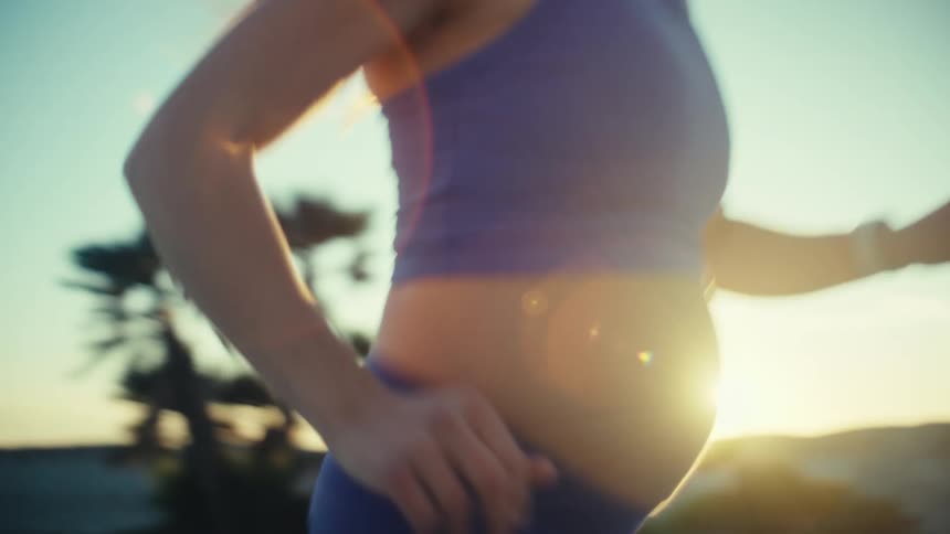 A view from the front shows a pregnant woman wearing a blue sports bra and blue leggings as she runs in an outdoor area with palm trees during sunset