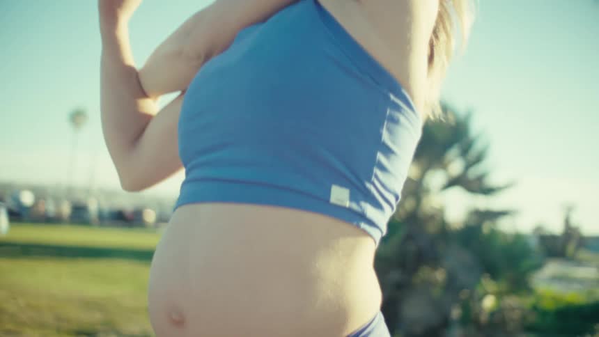 A pregnant woman wearing a blue sports top and leggings stretches her arms while exercising in a sunny outdoor park with palm trees and a bright clear sky in the background during a daytime fitness session