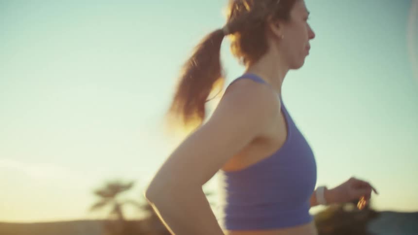 A pregnant woman in blue athletic wear jogs along a coastal path during a golden sunsetthe camera tracks her from the front before panning around to show her running toward a beachside building