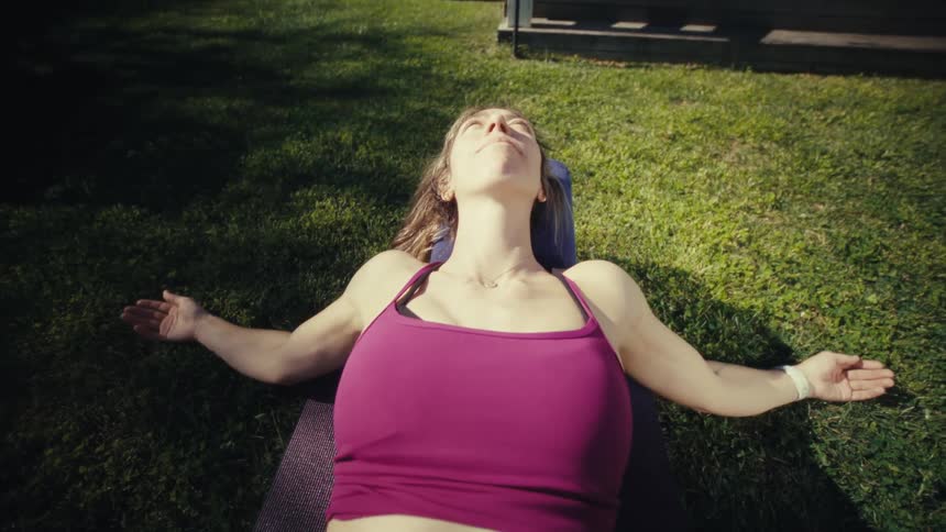 A pregnant woman lying and relaxing on a yoga mat on a lawn under bright sunlight