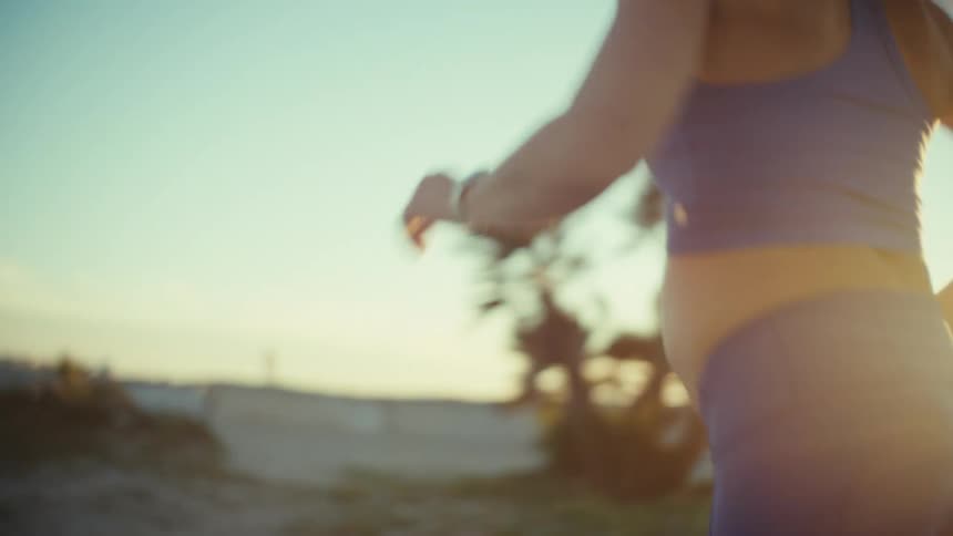 A woman with a ponytail wears blue athletic gear while jogging along a sandy coastal path during a bright sunset