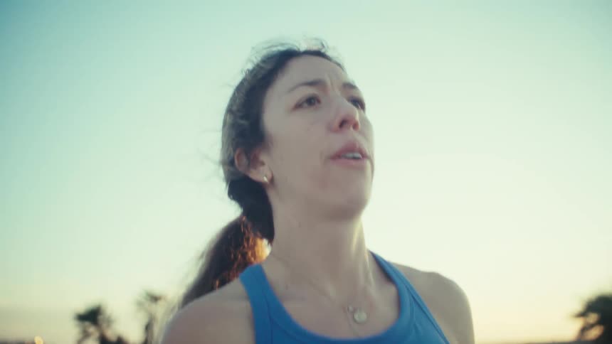 A woman in a blue tank top runs along a coastal path during a bright sunny dayas she slows to a stop she breathes heavily while the sunlight glows behind her and the palm trees