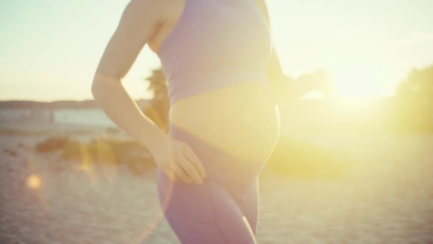 A pregnant woman wearing a purple sports bra and matching leggings runs along a sandy beach during a bright sunset with palm trees and soft lens flares in the background