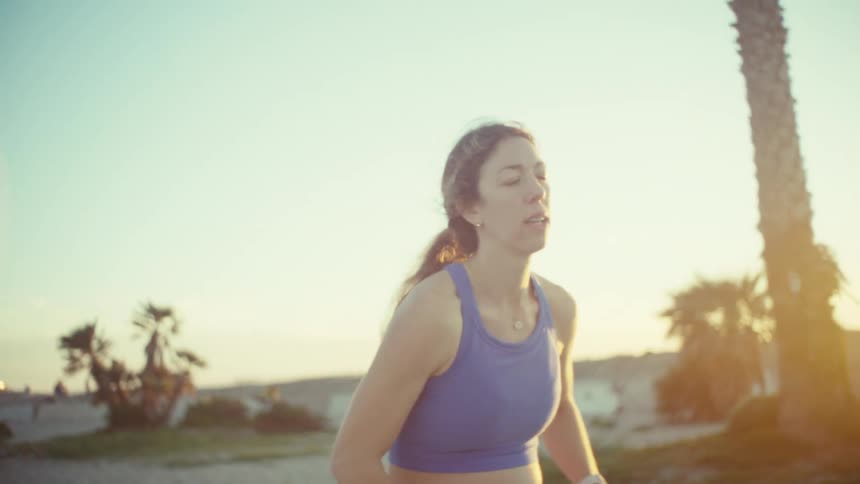A woman wearing a purple sports top and blue leggings stands outdoors on a beach during sunset and raises her arms toward the sky while palm trees and a clear horizon are visible in the background