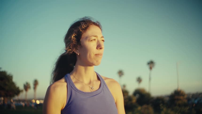 A woman wearing a blue sports top stands outdoors during sunset and removes her sunglasses while looking into the distance with palm trees in the background