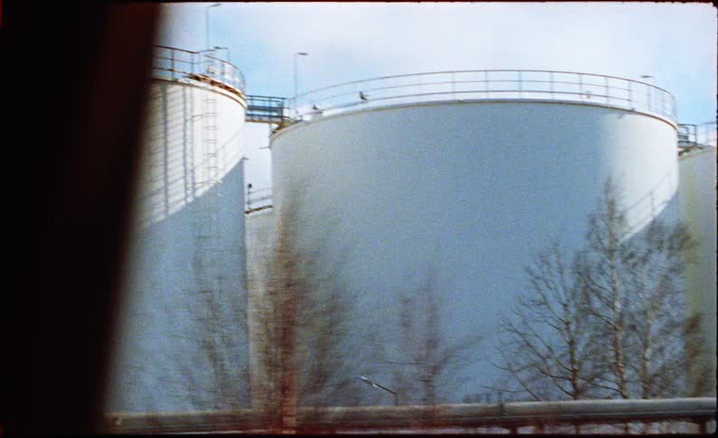 A moving camera captures large industrial storage tanks and complex piping systems under a bright cloudy sky