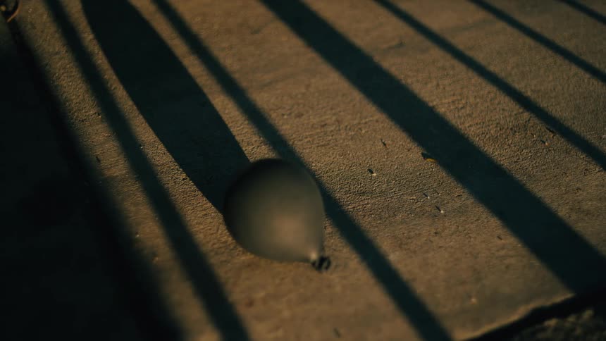 Long dark shadows from a metal fence stretch across a concrete ground where a single black balloon lies still in the dim light