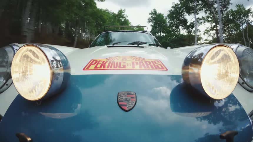 Lowangle closeup of a blueandwhite vintage porsche with its headlights blazing the porsche crest on the hood and a bright peking to paris rally decal above it as it speeds past a treelined road