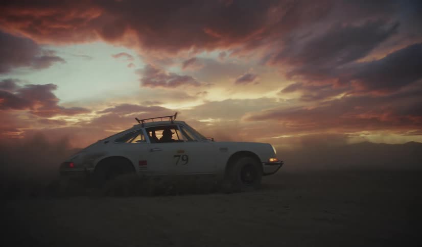 A white vintage racing car with a roof rack and the number seventy nine on the side drives through a flat desert landscape at sunset kicking up a thick cloud of dust under a dramatic sky with orange and purple clouds