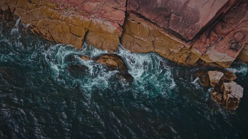 An aerial view shows powerful dark ocean waves repeatedly breaking against a rocky colorful shore