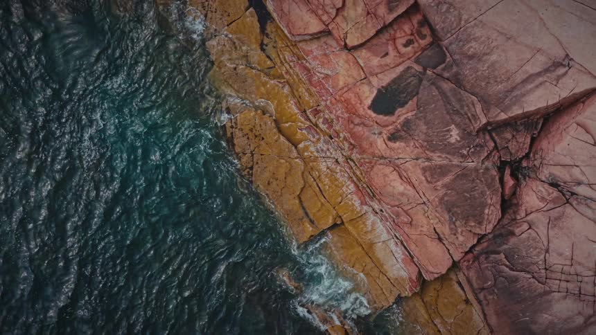 Dark ocean water meets a vibrant red and yellow textured rock formation in a serene overhead view