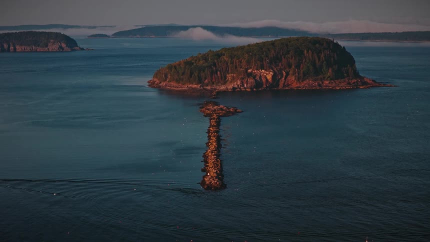 An aerial long shot captures a boat moving across a calm bay with a tree covered island and rock formation
