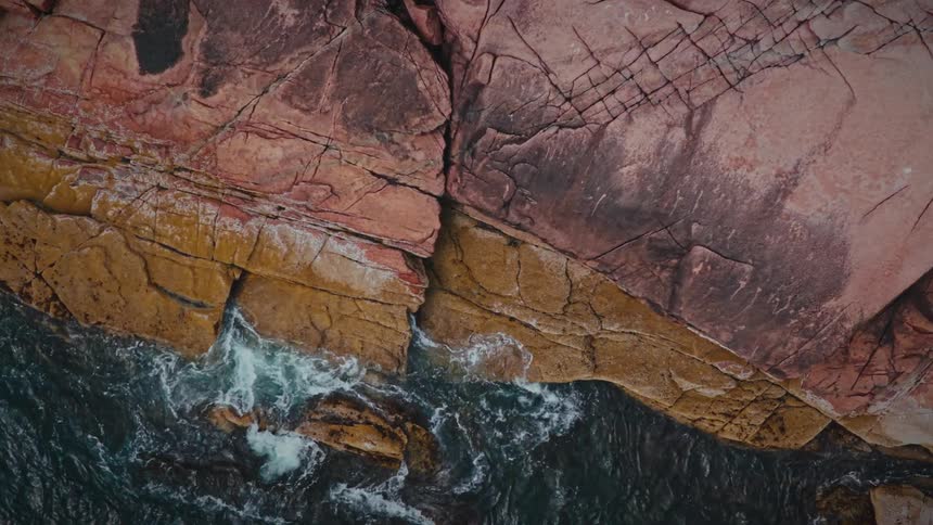 Waves crash rhythmically against multi layered textured rock formations along a rugged coastline