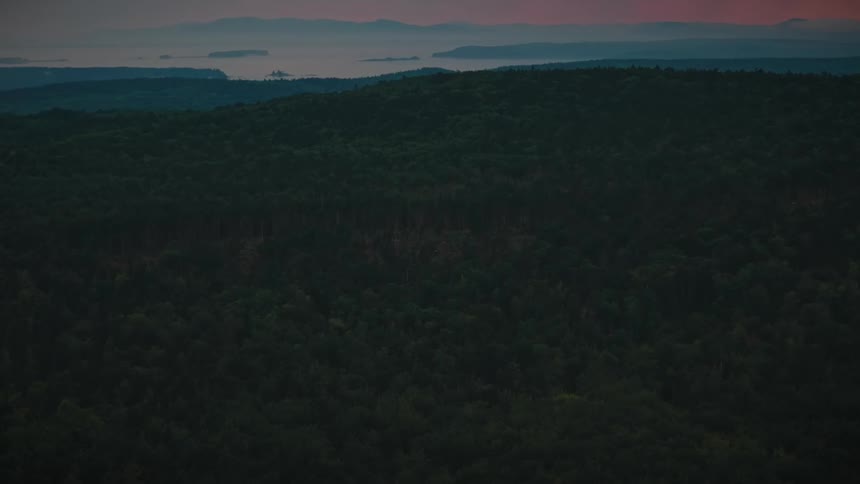 A high angle view shows a dense green forest covering rolling hills under a dark twilight sky with a faint orange glow on the distant horizon over a calm body of water
