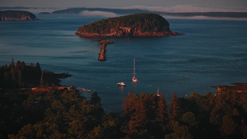 An aerial view shows a tranquil bay with moored boats a causeway leading to a forested island and misty coastal scenery