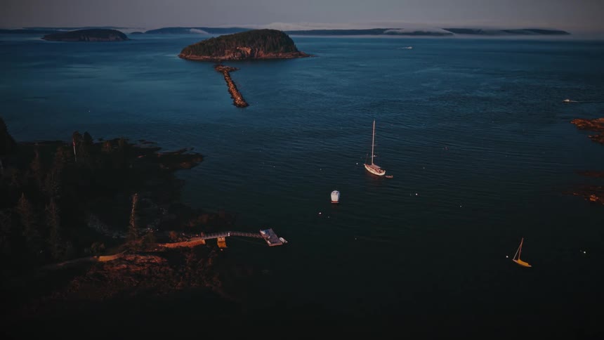 Aerial view of a sailboat and a small motorboat anchored in deep blue water beside a narrow rocky breakwater that leads to a small treecovered island with low islands on the distant horizon