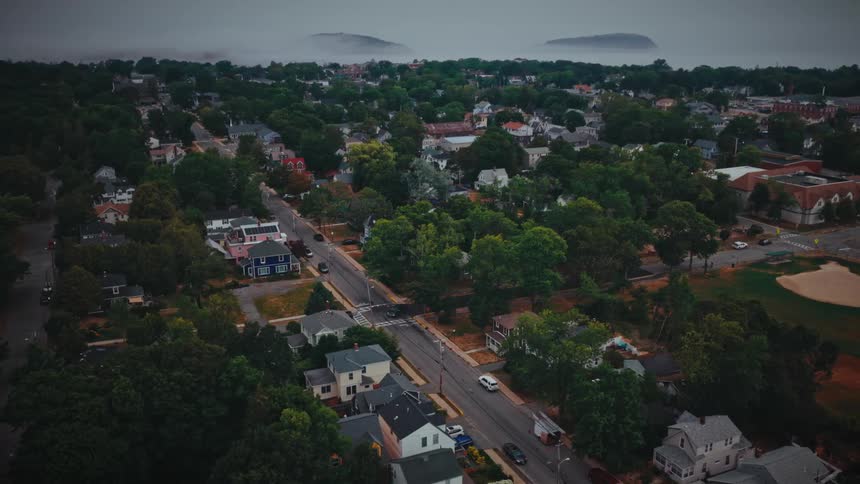 A high angle view shows a town with many houses and green trees along a straight road leading toward a foggy coast with small islands in the distance under a gray sky