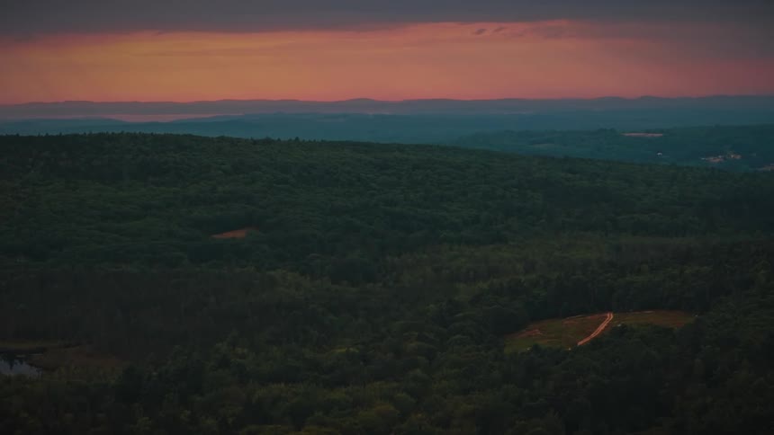 An expansive aerial view captures a dense forest under a colorful sunset sky with distant mountains