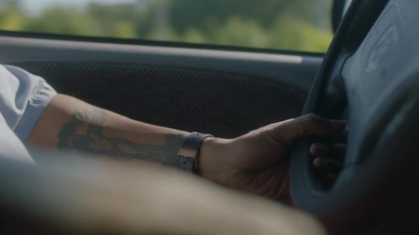 A young man with a tattooed left arm is driving a car during the daytime