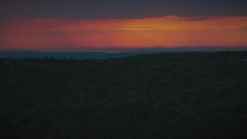 A wide landscape view shows a dense green forest under a vibrant orange and purple sunset sky where layers of distant mountains sit on the horizon below heavy clouds