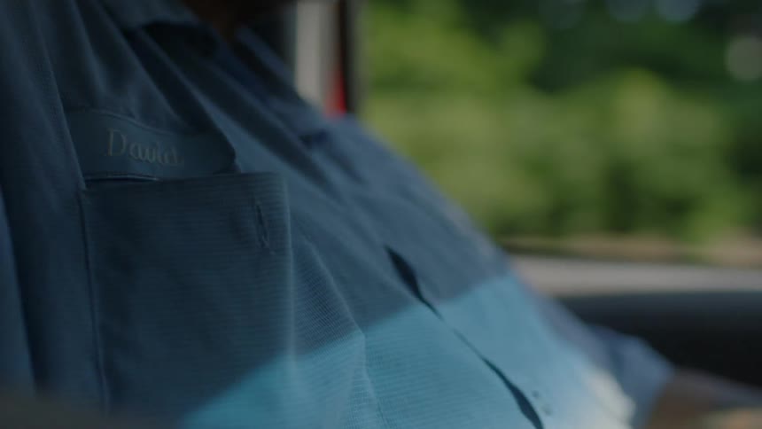 A person wearing a mechanics work shirt is driving a vehicle during the day