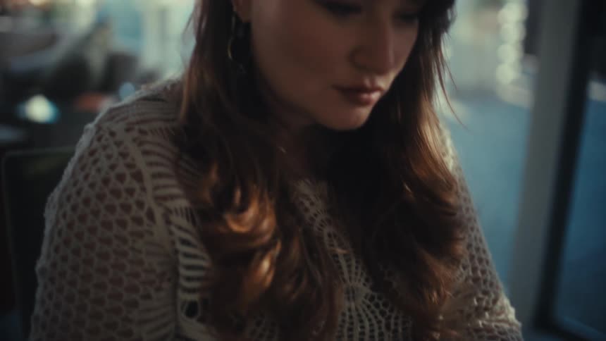 A woman wearing a white lace top and gold rings sits in a dimly lit room typing on a laptop computer with a focused expression while soft light reflects on her face