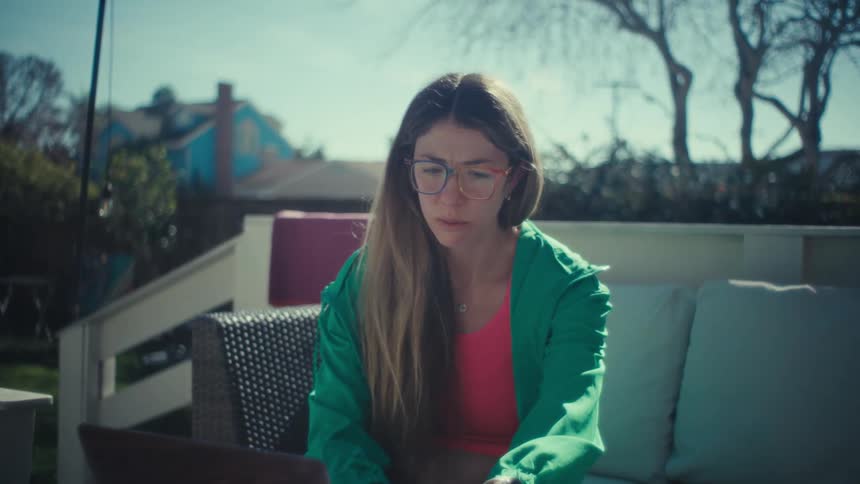 A young woman studies a book and types on a laptop while sitting outdoors