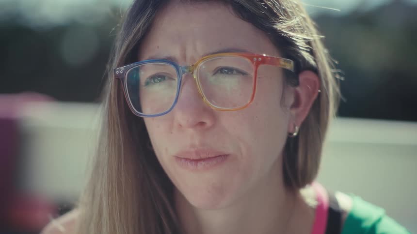 A woman wearing colorful glasses and a green jacket sits outdoors and looks with a thoughtful expression in the bright sunlight